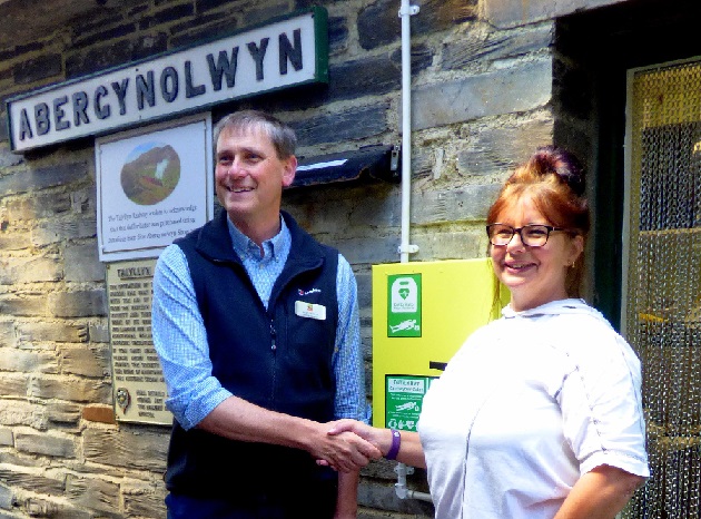Two people shake hands in front of the defibrillator mounted on a stone wall.