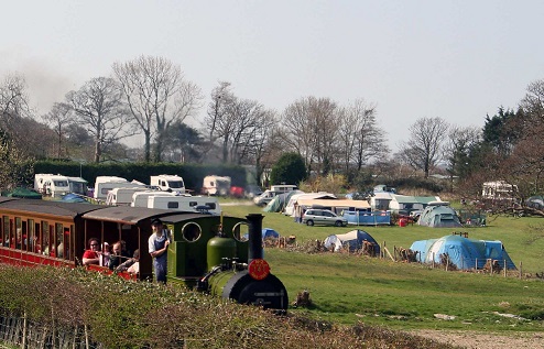 A steam train passing by the caravan park with tents and mobile homes in the background.