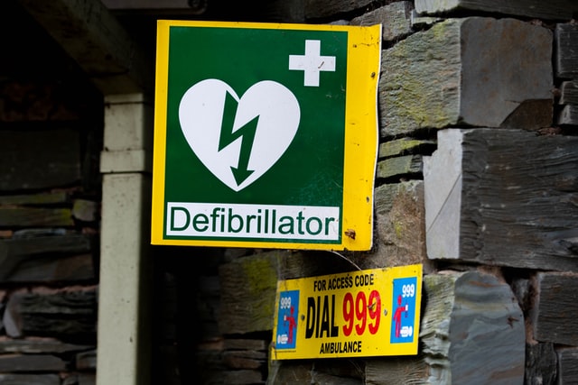 A yellow defibrillator mounted to the wall of the slate toilet building.