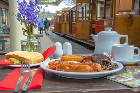 A cooked breakfast on a table outside in front of a steam hauled train.
