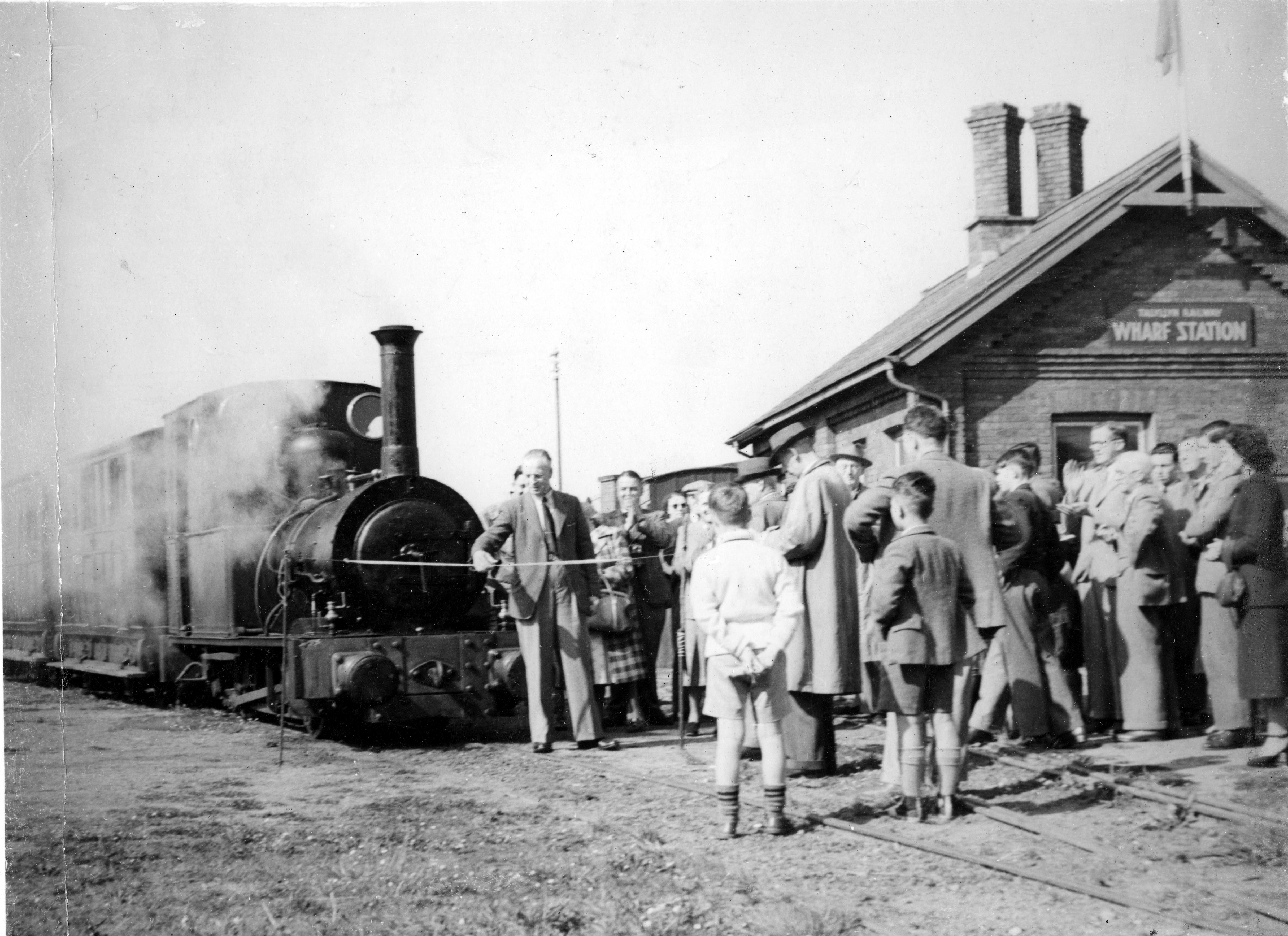 Black and white image of a man curring a ribbon in front of a narrow gauge engine.