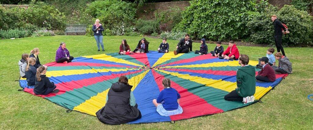 A group of children and young adults sat on a multicoloured fabric circle on a lawn.