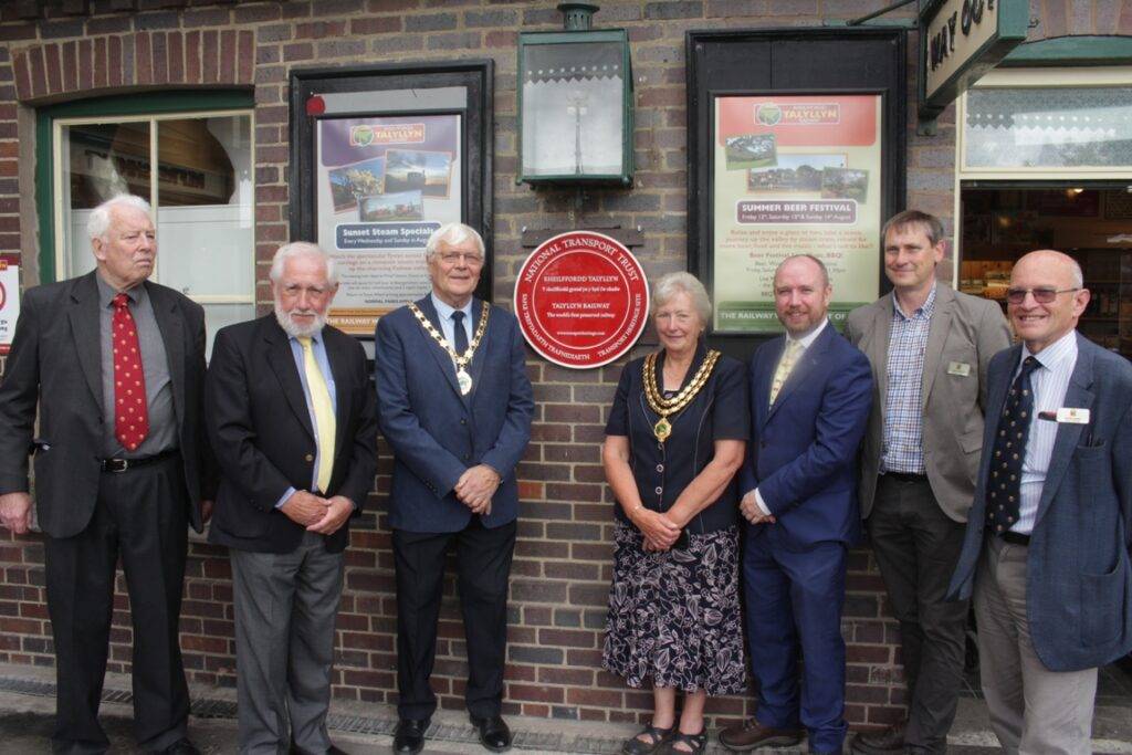 Seven people stood on a railway station platform with a round red plaque on the wall behind them.