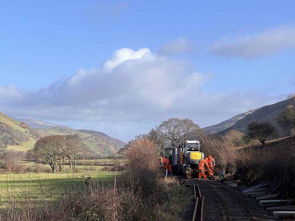 A lenght of railway line with th etrack removed. There are people, a digger and a train in the distance.